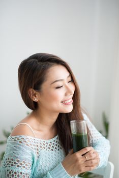 Smiling young asian woman drinking green fresh vegetable juice or smoothie from glass at home