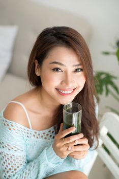 Pretty young asian woman holding green fresh vegetable juice or smoothie from glass at home