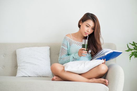 Pretty young asian woman drinking green fresh vegetable juice or smoothie from glass at home