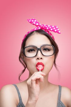 Portrait of beautiful asian woman eating heart shape lollipop, dressed and makeup in pin-up style on pink background.