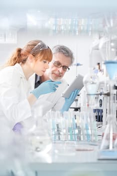 Health care researchers working in life scientific laboratory. Young female research scientist and senior male supervisor looking focused at tablet computer screen evaluating and analyzing study data.