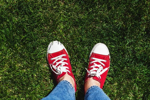 Legs in old red sneakers on green grass. View from above. The concept of youth, spring and freedom.