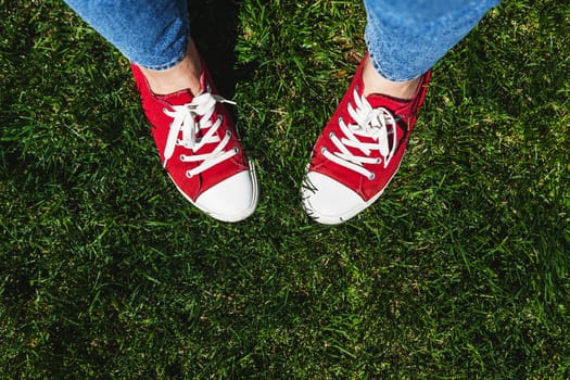 Legs in old red sneakers on green grass. View from above. The concept of youth, spring and freedom.