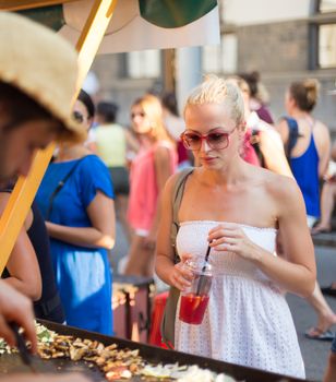 Beautiful blonde caucasian lady wearing white summer dress buying freshly prepared meal at a local street food festival. Urban international kitchen event in Ljubljana, Slovenia, in summertime.
