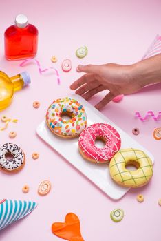 Party. Hand holding plate of colourful sugary round glazed donuts and bottles of drinks on pink background.