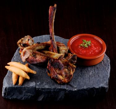Delicious Roasted Lamb Ribs with Bread Sticks and Tomato Sauce on Stone Plate closeup on Dark Wooden background