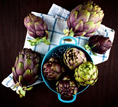Stack of Perfect Raw Artichokes in Blue Colander closeup on Checkered Napkin closeup on Dark Wooden background. Top View
