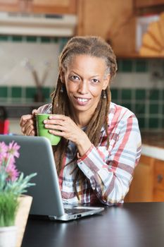 Single adult woman with dreadlocks in kitchen holding green mug in front of laptop