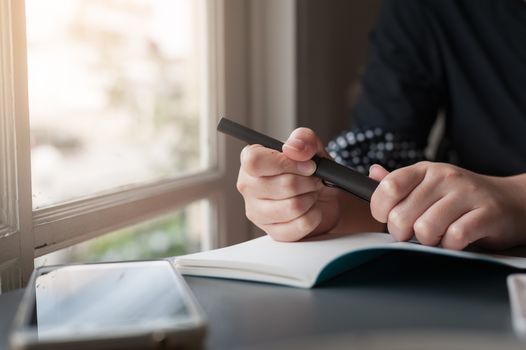 Woman hand holding pen while writing on small notebook beside window. Freelance journalist working at home concept.