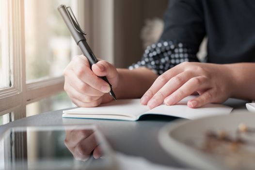 Woman hand holding pen while writing on small notebook beside window. Freelance journalist working at home concept.