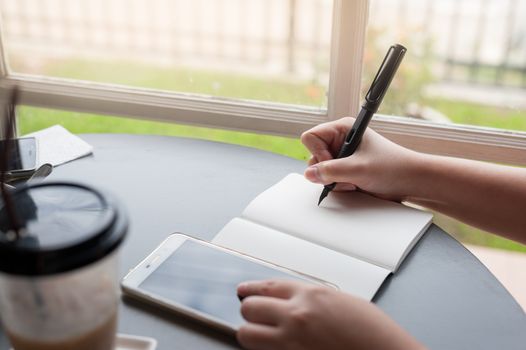 Woman hand writing a note on notebook while another hand touching on smartphone screen on table beside window. Working from anywhere concept