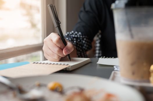 Woman hand holding pen while writing on small notebook beside window. Freelance journalist working at home concept.