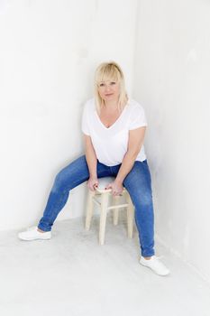 Blond woman in white and blue jeans sitting on the stool inside empty room