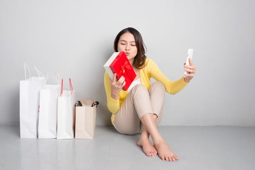 Young asian woman sitting besides row of shopping bags holding red gift box taking sellfie