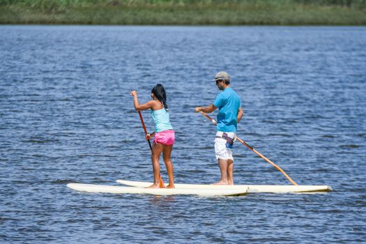 Man and woman stand up paddleboarding on lake. Young couple are doing watersport on lake. Male and female tourists are in swimwear during summer vacation.