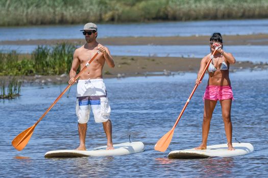 Man and woman stand up paddleboarding on lake. Young couple are doing watersport on lake. Male and female tourists are in swimwear during summer vacation.