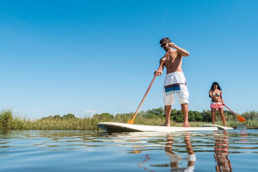 Man and woman stand up paddleboarding on lake. Young couple are doing watersport on lake. Male and female tourists are in swimwear during summer vacation.