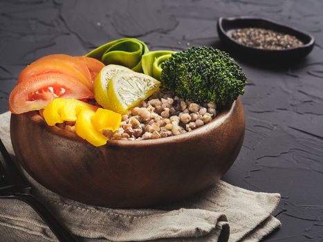 Close up view of vegetarian buddha bowl with green buckwheat, broccoli, avocado, tomatoes and yellow sweet pepper paprika on dark concrete background with copy space