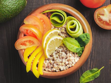 Top view of vegetarian buddha bowl with green buckwheat, spinach, avocado, tomatoes and yellow sweet pepper paprika on dark wooden table with copy space
