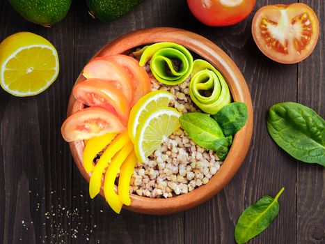 Top view of vegetarian buddha bowl with green buckwheat, spinach, avocado, tomatoes and yellow sweet pepper paprika on dark wooden table with copy space