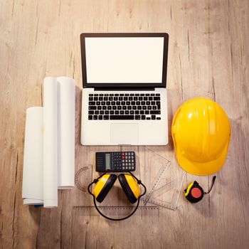 Top view of a architectonic workspace with laptop and yellow helmet. Different accessories, plans and cup of coffee is on the table. 