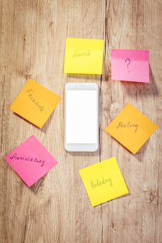 Top view of a white smartphone with blank screen and many adhesive notes with brainstorm on wooden table.