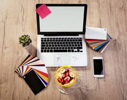 Top view of a designer workspace with laptop and pallete color. Different office accessories, smartphone and cup of fruit salad is on the table. 