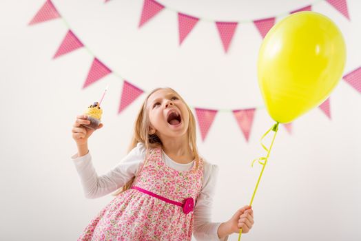 Beautiful cheerful little girl holding balloon and small birthday cake with candle.