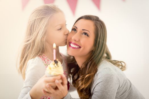 Cute little girl giving small cupcake with candle for her smiling mom. Selective focus. Focus on background, on mom.