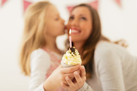 Cute little girl giving small cupcake with candle for her smiling mom. Selective focus. Focus on foreground, on cake.