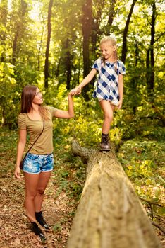 Beautiful young mother and her daughter having fun in the forest. Mother holding girl who walking over fallen tree.