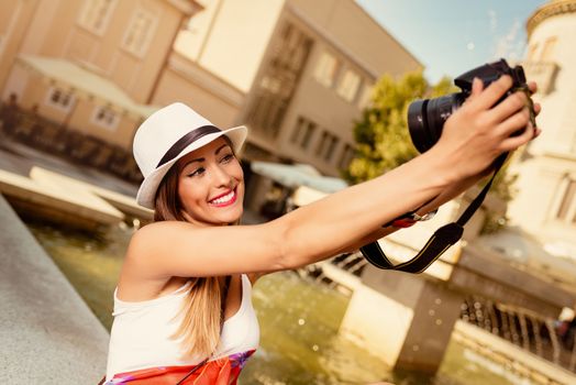 Beautiful young smiling woman with hat taking selfie in the city square next to the fountain.