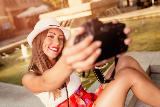 Beautiful young smiling woman with hat taking selfie in the city square next to the fountain.