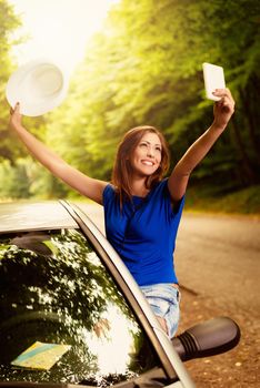 Cheerful woman on summer travel vacation leaning out of a car window. She is holding hat with arms raised and taking selfie.
