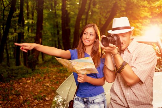 Young travelers standing before a car in the forest. Girl holding map and pointing destination. Guy taking a photo with digital camera.