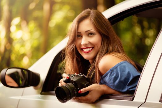 Beautiful young woman photographer sitting in car at the forest, holding digital camera and pensive looking away.