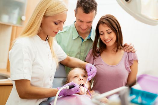 Happy family at visit in the dentist office. Female dentist checking teeth the little girl, her parents standing next to her.