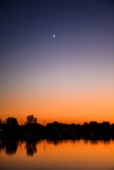 A crescent moon is seen at sunset over a lake