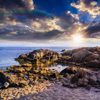 sea coast with boulders sand and seaweed with rainbow at sunset