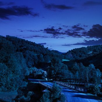 asphalt road going through the bridge off into mountains on the left, passes through the green shaded forest at night