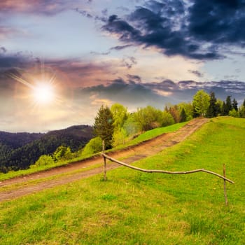 summer landscape. fence near the meadow path going up on the hillside. forest in fog on the mountain at sunset