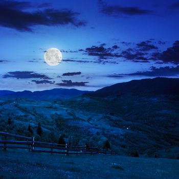 summer landscape. fence near the meadow on hillside. forest in fog on the mountain at night in moon light