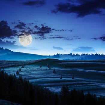 Stack of hay on a green meadow in the mountains at night in moon light under a blue summer sky