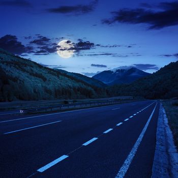 stright asphalt road in mountains passes through the green shaded forest at night in moon light