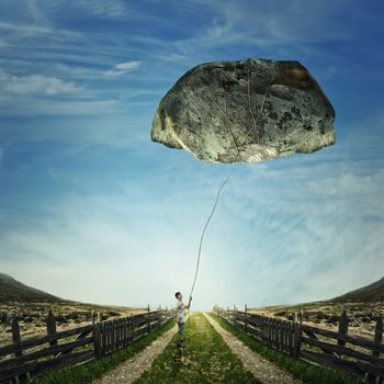 Surrealistic image as a young boy stand on a country road, holding a rope bound around a huge stone as a playing kite. Life pressure, stress and hard determination concept.
