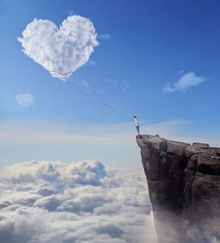 Imaginary view with a boy on the edge of a cliff, trying to catch a heart shaped cloud with a long rope. Follow your heart concept.