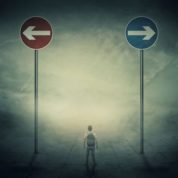 Surrealistic image as a man, with a bag in his back, stand in a foggy street in front of two huge road signs that show the left and right directions. Strategic journey, way of life and intuition concept.