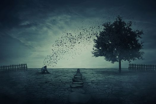 Young man sit alone on the cracked ground, in the middle of his farm garden, in front of a magic tree that cast its leaves in the wind. Energy changing concept, between people and nature.