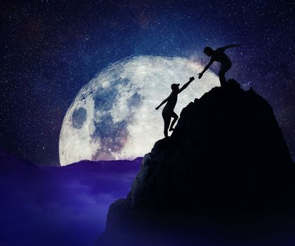 Man giving helping hand to friend to climb mountain rock cliff. Teamwork and trust concept in risky situation. Full moon night background. 