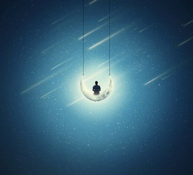 Surreal background with a lonely boy sitting on a crescent moon, as a swing, over a starry night sky 